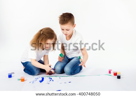 Two pretty child friends boy and girl in white shirts and blue jeans, trendy hair style, barefoot, drawing pictures on white sheet of paper by paints isolated on white. Studio shot.