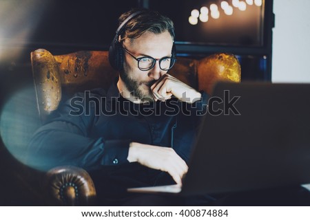 Photo bearded businessman wearing glasses relaxing modern loft office.Man sitting in vintage chair,listening music laptop night.Using contemporary notebook, blurred background.Horizontal,film effect.  Royalty-Free Stock Photo #400874884
