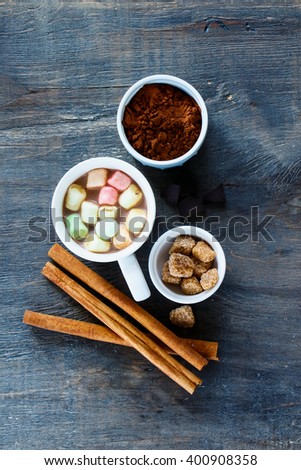 White cup of hot chocolate with melting marshmallows and cinnamon sticks over vintage kitchen table, top view. Dark rustic style.