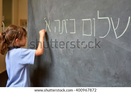 Young Israeli Jewish girl (Age 5-6) writes Hello First Grade greetings in Hebrew (Shalom Kita Alef) on chalkboard in Israeli primary school at the beginning of the school year. Real people.Copy space Royalty-Free Stock Photo #400967872