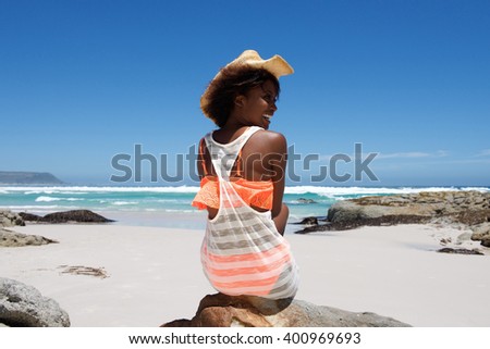 Rear view portrait of cheerful young woman with hat sitting by the beach