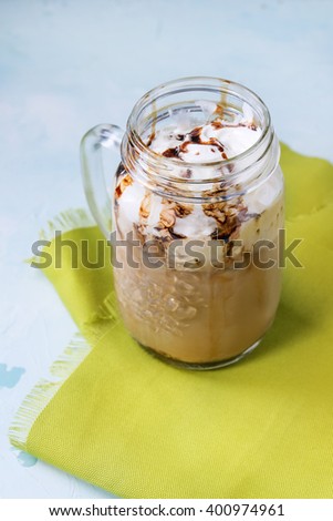 Glass mason jar with ice coffee with whipped cream, ice cream and chocolate sauce, served with coffee beans and ice cubes on green textile napkin over light blue textured background.