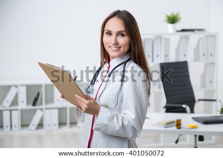 Smiling female doctor standing at table and making notes. Office at background. Concept of work.