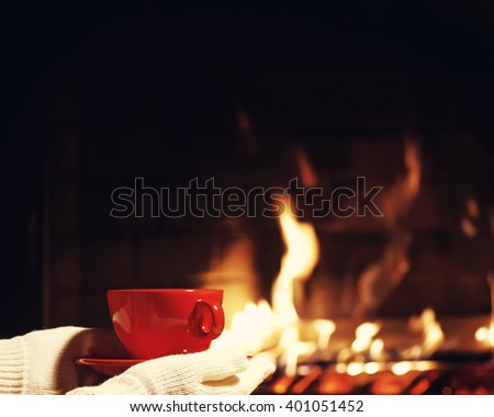 Red cup of tea or coffee in hands dressed in warm mittens near  fireplace. Winter and Christmas holiday concept. Photo with retro filter effect.