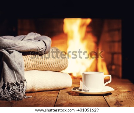 White cup of tea and warm woolen things near fireplace on wooden table. Winter and Christmas holiday concept. Photo with retro filter effect.
