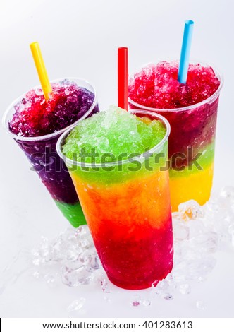 Still Life Close Up of Colorful Rainbow Layered Frozen Fruit Slush Drinks Arranged on Ice Covered White Surface in Plastic Take Away Cups with Drinking Straws - Trio of Refreshing Granitas Royalty-Free Stock Photo #401283613