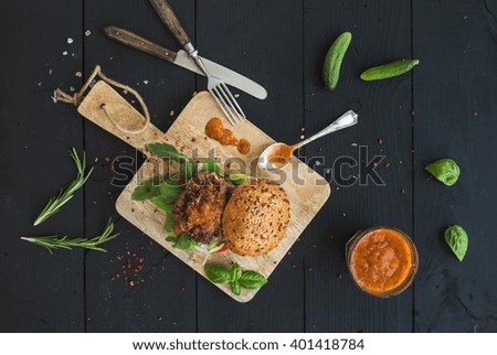 Fresh homemade burger on dark serving board with spicy tomato sauce, sea salt and herbs over dark wooden background. Top view