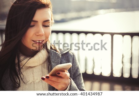 Attractive young brunette girl with long brown hair and friendly smile using a modern smart phone outdoors at bright spring day. 