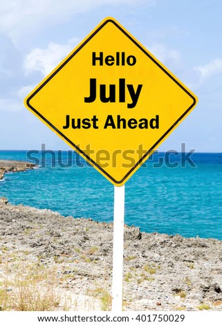 Yellow road sign with a Hello July Just Ahead concept against a coastal setting with a partly cloudy sky background.
