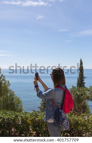 Adult brunette with pink backpack making photo with phone while standing against of beautiful water scape. Copy space.
