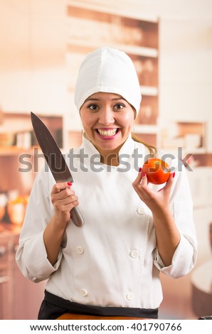 Woman chef holding up large metal knife and tomato while smiling to camera
