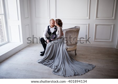 the couple are facing each other and embracing on the vintage couch in a white room, the bride in a gray dress with lace, groom with red beard in black trousers, white shirt and black vest Royalty-Free Stock Photo #401894134