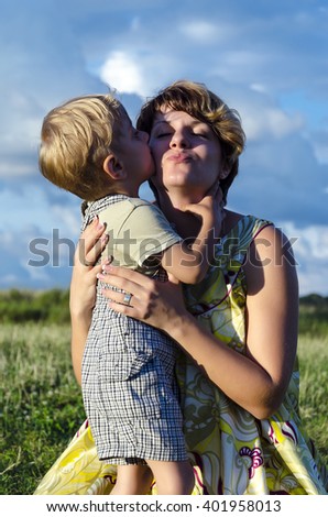 Portrait of happy mom kissing and shake son on green summer garden. Cute  mother embracing baby boy looking at camera