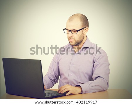 Busy man with beard in glasses thinking over laptop with  on the table. Isolated on gray background