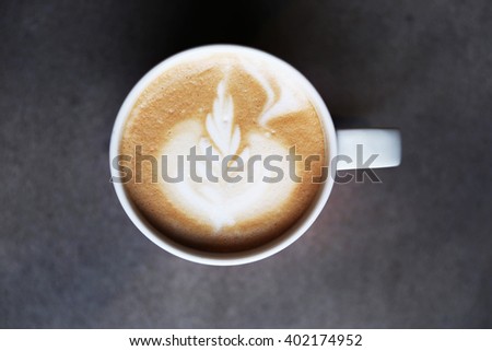 Cup of tasty latte is standing on the grey textured table. There is nice picture is painted on the top of the latte. Latte is in the white big cup.
