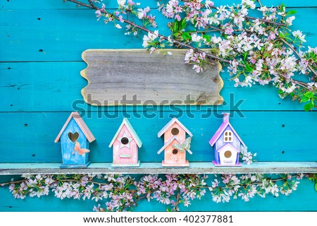 Blank wood  sign hanging over colorful birdhouses with butterfly on shelf by spring tree flowers on antique rustic teal blue wooden background; pink, purple, orange birdhouses