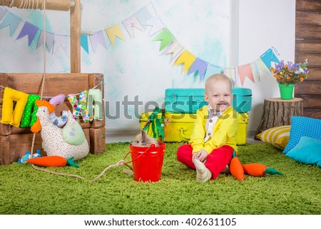 Little boy in bright clothes in the bright studio is played with the Easter bunny in the grass on the background of the check boxes and suitcases