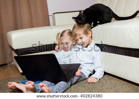 two happy little baby boy and a fair-skinned blonde girl (brother and sister) sitting on the floor with a laptop and watching cartoons on the computer, a cat on a sofa ironed (fins) on kid