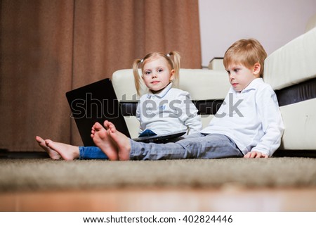 two little blond baby boy and a fair-skinned girl (brother and sister) sitting on the floor with a laptop, watch cartoons and play computer games