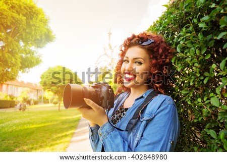 Outdoors portrait of fashionable young curly redhead female photographer holding digital camera, smiling, standing in park on sunny spring day, wearing denim shirt. Horizontal, vibrant, retouched.