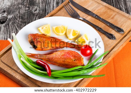 Red Sea Bass on awhite dish on an old wooden table with green spring onions, lemon slices and cherry tomato on the background, horizontal close-up