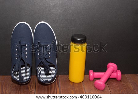 Blue fitness shoes, orange juice and pink weights on wooden floor in front of black desk