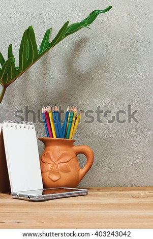 Pencil in earthenware vases, mobile phone and notebook on a wooden table under a foliage plant.