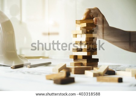 Hand of engineer playing a blocks wood game (jenga) on blueprint or architectural project vintage tone. Double exposure with back side of business man and flight control tower. Royalty-Free Stock Photo #403318588