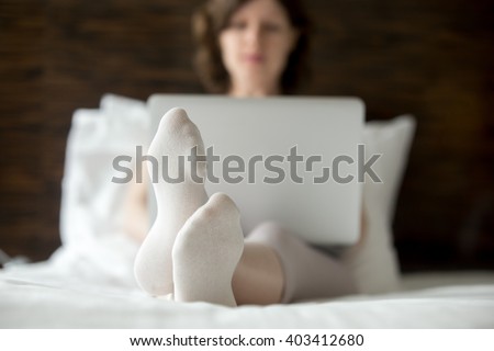 Beautiful young woman sitting on bed using laptop in modern room. Caucasian model relaxing with computer indoors, surfing internet, shopping on-line. Close-up of feet