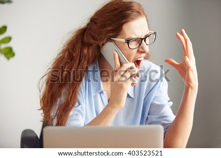 Headshot of a female boss shouting with anger on cell phone while sitting at desk in office. Portrait of an angry businesswoman screaming on mobile phone. Negative human emotions, facial expressions Royalty-Free Stock Photo #403523521