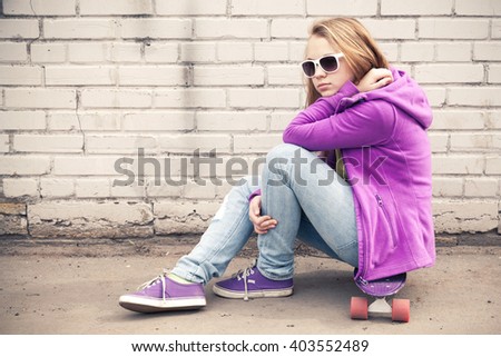 Blond teenage girl in jeans and sunglasses sits on her skateboard near white brick wall, photo with warm retro tonal correction effect, old style filter