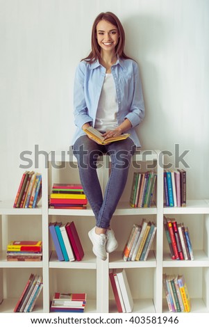 Beautiful young girl in jeans is holding a book overhead, looking at camera and smiling, sitting on the top of bookcase