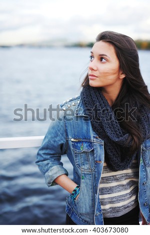 beautiful brunette girl in a denim jacket and a scarf standing near a railing near the water
