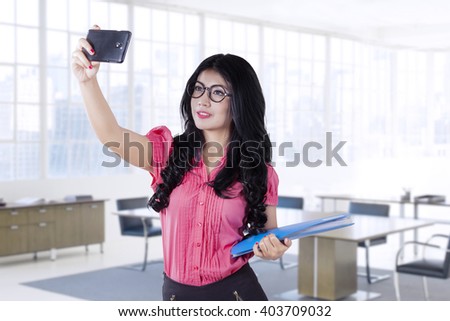 Happy businesswoman with long hair, taking selfie picture with smartphone in the office