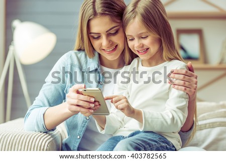 Beautiful young mom and her little daughter are using a smartphone and smiling while sitting on sofa at home Royalty-Free Stock Photo #403782565