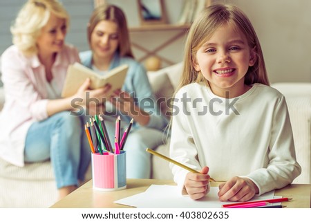 Little girl is drawing and smiling, in the background her mom and granny are reading a book while sitting on sofa at home
