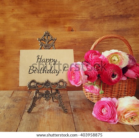 pink flowers in the basket next to card with phrase: happy birthday, on wooden table.
