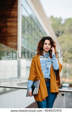 Smiling curly haired woman with laptop in knitted poncho talking cell phone. Unfocused urban background