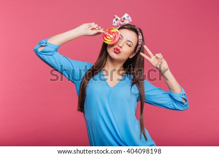 Cute playful young woman covered her eye with lollipop and showing peace sign over pink background