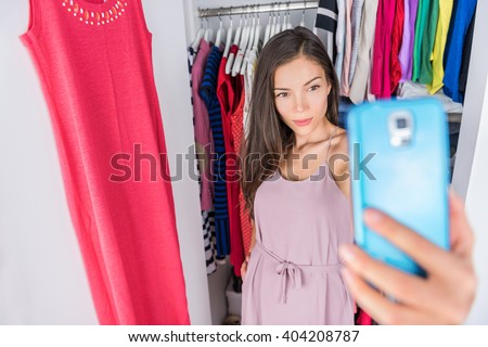 Smart phone selfie Asian woman in clothes closet of home bedroom or  store dressing room next to clothing rack. Shopping girl taking a photo of her outfit using smartphone fashion app. Social media.