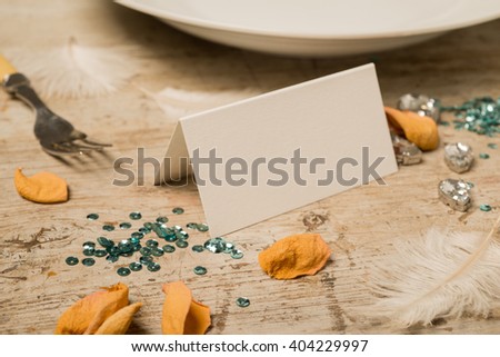 Empty place card facing right surrounded by green sequins, feathers, fake gemstones, dinner fork and plate, and flower petals on a wooden surface for copy space.