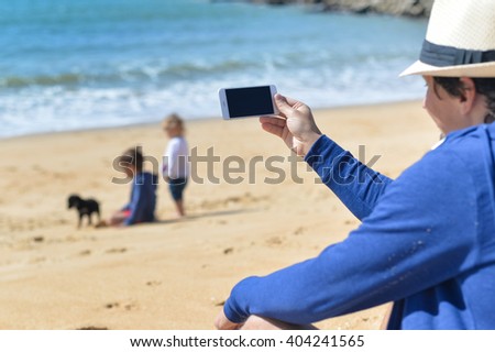 Back view shot of man holding smartphone photographing selfie or beach background outdoors. Happy vacation travel concepts