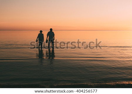 Silhouette of adult couple which costs in the sea and holds in hand footwear against a sunset.