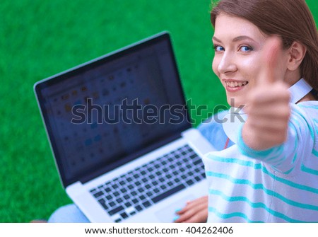 Young woman with laptop sitting on green grass and showing ok