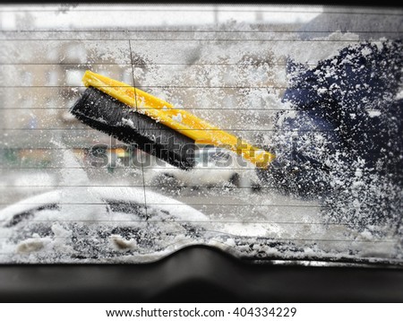 Cleaning of car window of snow with use of yellow brush. Look from within salon. Through muddy wet glass are visible winter street with  building,  car, part of  spare wheel.  