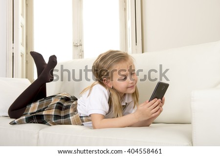sweet cute and beautiful 6 or 7 years old female child with blond hair in school uniform lying on home sofa couch using internet app on mobile phone playing online game looking happy and relaxed