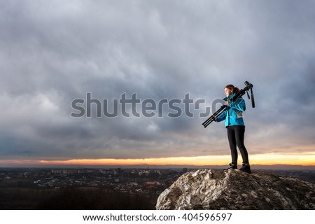 Young attractive photographer is standing with her camera on tripod on the big rock at city overview point at the sunset. Girl is holding tripod on her shoulder. Blue evening sky in the background
