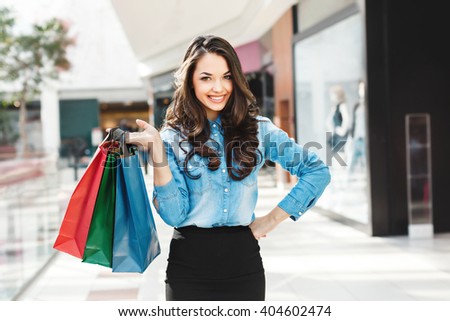 Beautiful girl in shopping mall, looking at camera, smiling and holding colorful shopping bags in one raised hand. Other hand on waist. Wearing blue jeans blouse and black skirt, nice make up. Indoor