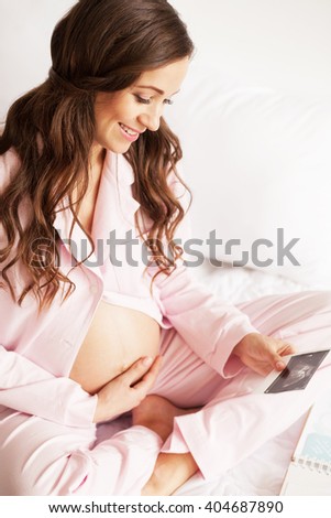 Lovely smiling pregnant woman sitting in bed and looking at ultrasonic scan of baby