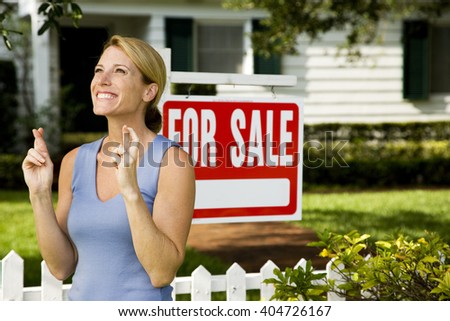 Woman standing by a for sale sign outside a family house, fingers crossed